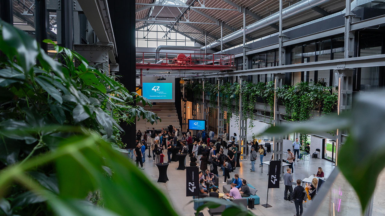 Das Bild zeigt eine Halle, links und rechts hängen in der ersten Etage Pflanzen. Im Erdgeschoss sind viele Personen und Werbebanner von 42Heilbronn zu erkennen.