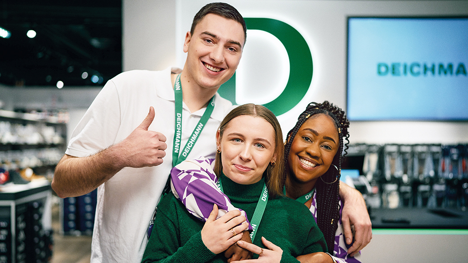 Drei Deichmann-Mitarbeitende stehen in einer Filiale und lächeln in die Kamera