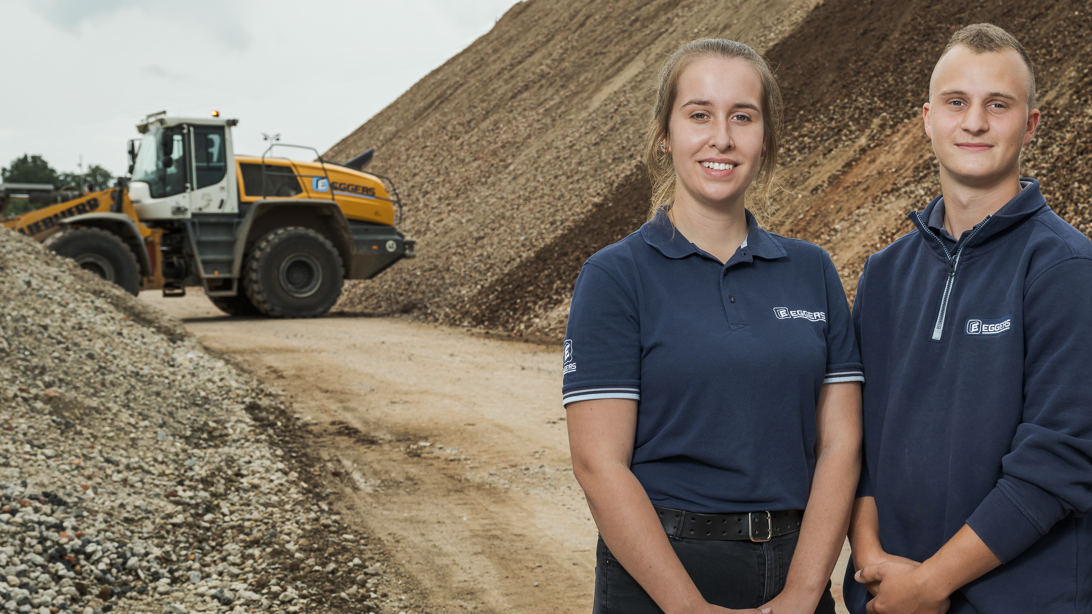 Zwei junge Mitarbeitende in Arbeitskleidung stehen auf Baustelle und ein Bagger steht im Hintergrund.