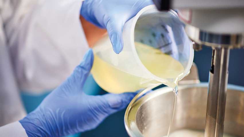 Mensch schüttet mit behandschuhten Händen Flüssigkeitn in einen Behälter