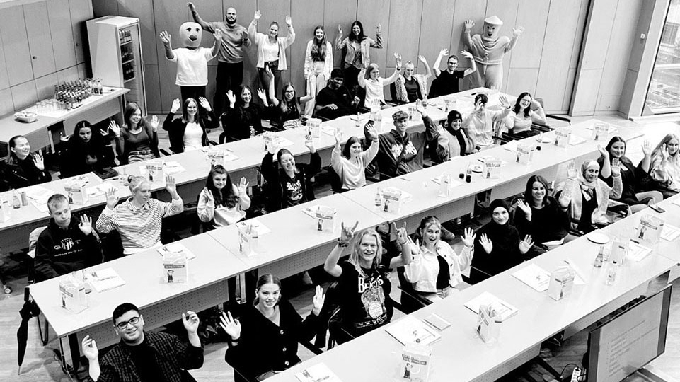 Eine große Gruppe von Menschen sitzt an langen Tischen in einem Saal und hebt lächelnd die Hände. Im Hintergrund stehen zwei Maskottchen.