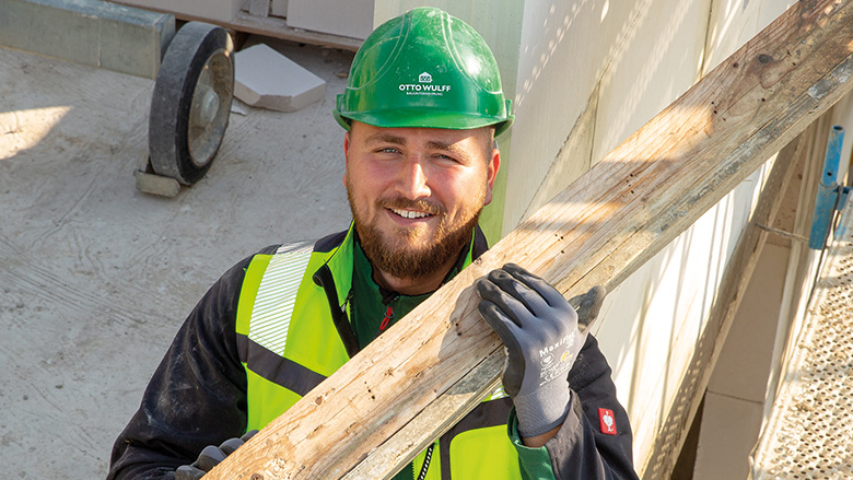 Mensch mit grünem Schutzhelm und einem Holzbalken über der Schulter lächelt in die Kamera