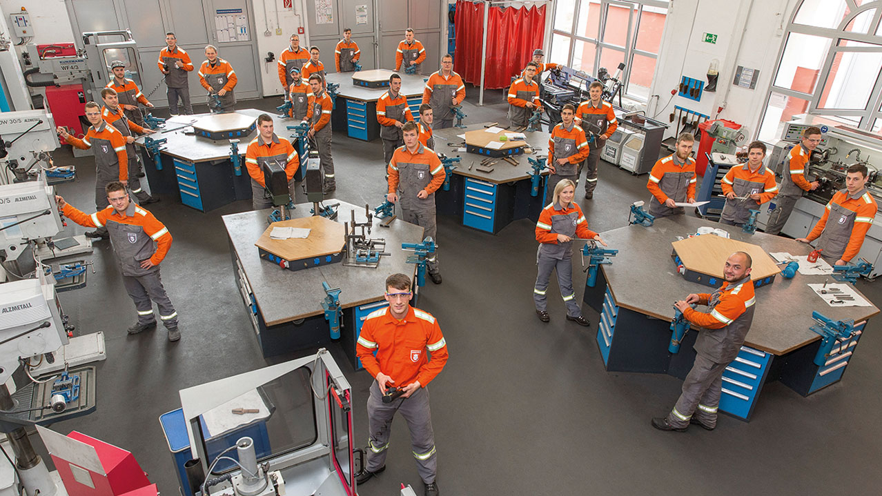 Mitarbeiter/innen in organge-grauer Arbeitskleidung stehen in einer Werkstatt und werden von oben fotografiert.