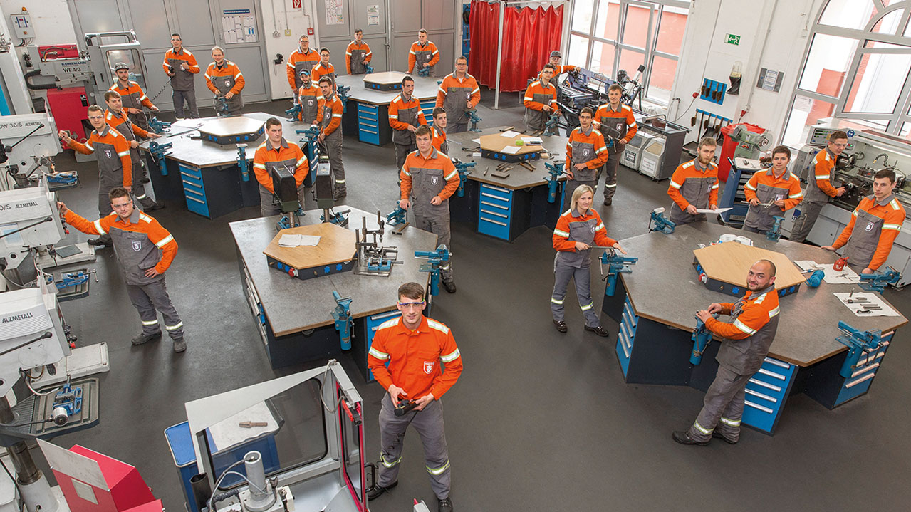 Mitarbeiter/innen in organge-grauer Arbeitskleidung stehen in einer Werkstatt und werden von oben fotografiert.