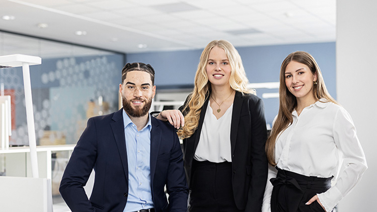 Ein junger Mann und zwei junge Frauen in Business Kleidung als Gruppe nebeneinander in einem modernen Büroraum.