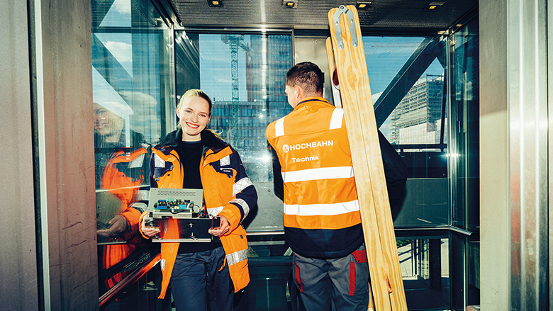 Zwei Mitarbeitende in Arbeitskleidung in einem gläsernen Aufzug