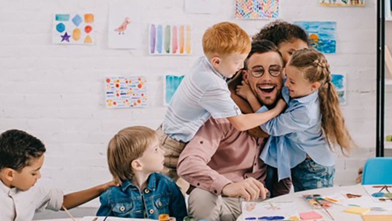 Eine junge Person sitzt an einem Tisch in einem Kindergarten. Kinder tollen um die Person herum.