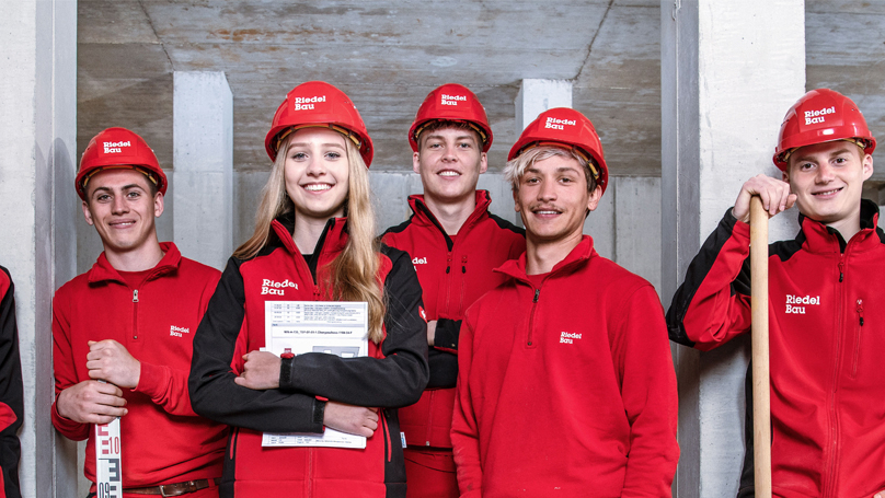 6 junge Mitarbeitende der Riedel Bau AG in roter Arbeitskleidung mit Helmen lächeln den Betrachter an.
