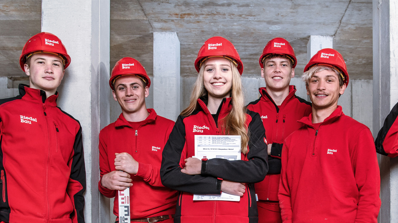 6 junge Mitarbeitende der Riedel Bau AG in roter Arbeitskleidung mit Helmen lächeln den Betrachter an.