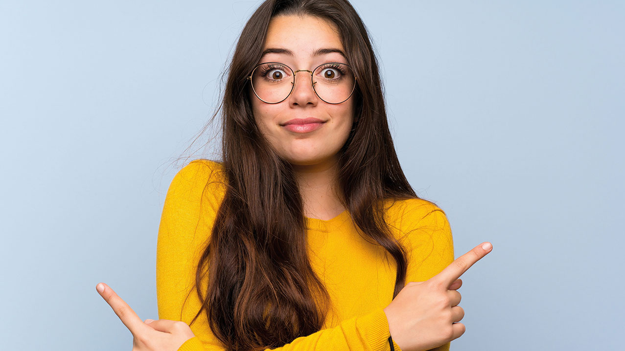 Dunkelhaariges Mädchen mit großer Brille schaut fragend. Ihre Arme sind überkreuzt und die Zeigefinder zeigen in entgegengesetzte Richtungen.