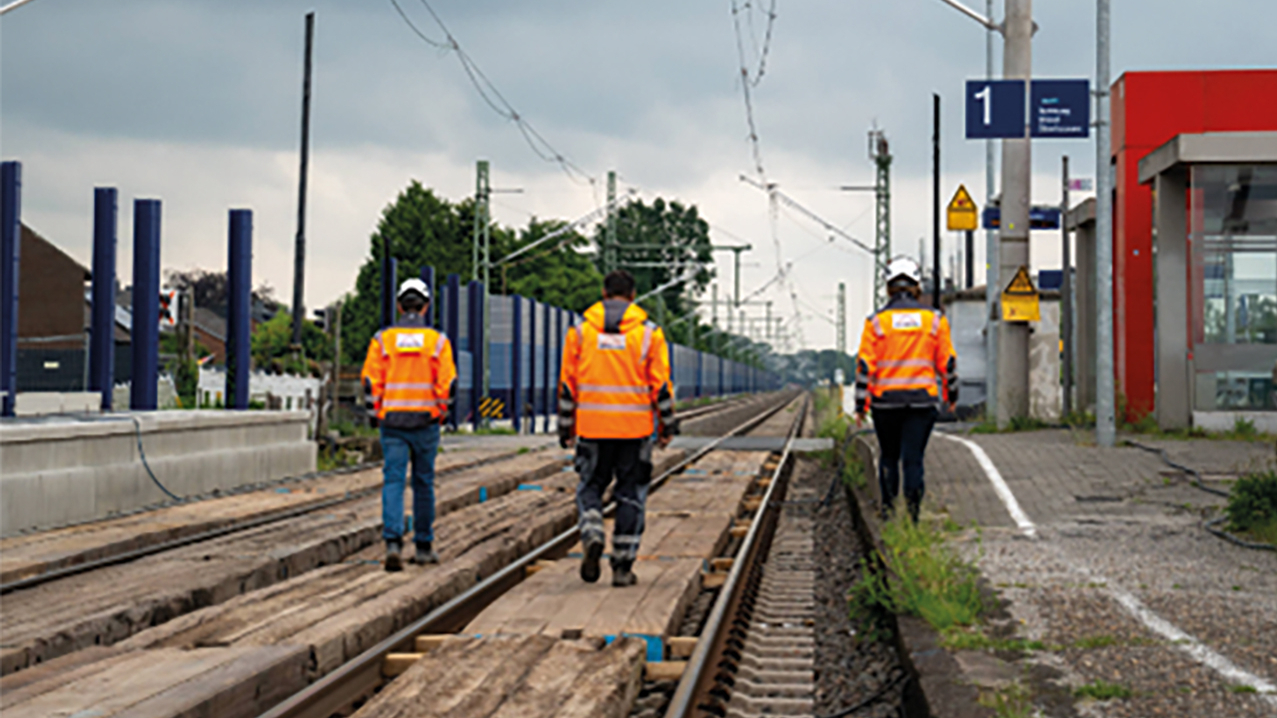 3 Personen in orangen Arbeitsjacken laufen auf Gleisen entlang.