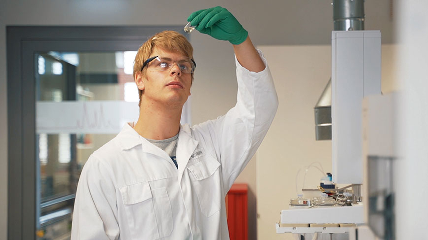 Person in weißem Kittel mit Schutzbrille untersucht eine Flüssigkeit im Labor