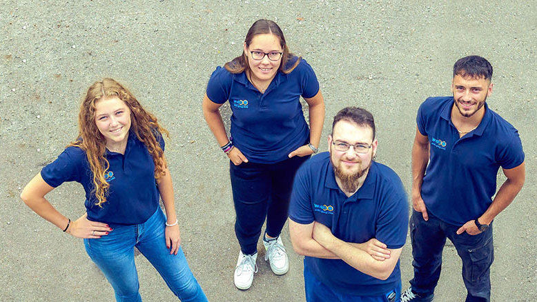 Vier Auszubildende in blauen Poloshirts schauen nach oben in die Kamera