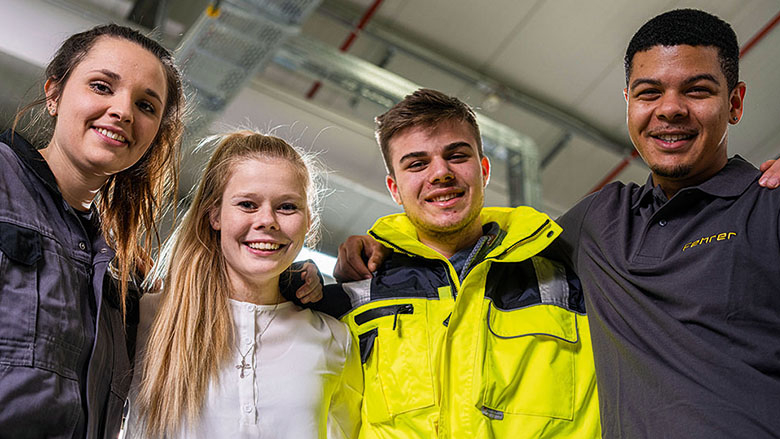 Zwei junge Frauen und zwei junge Männer die eng nebeneinander stehen und sich an den Schultern festhalten lächeln in die Kamera.