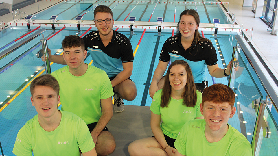 Sechs lächelnde Azubis vor dem Schwimmbecken