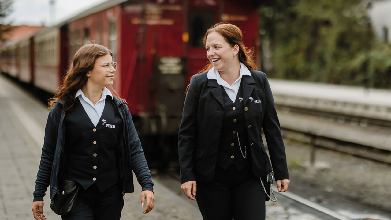 Zwei Schaffnerinnen laufen lächelnd vor einer harzer Schmalspurbahn auf dem Bahnsteig.