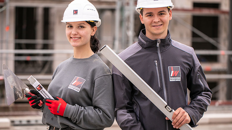 Zwei Mitarbeitende in Arbeitskleidung mit Zollstock, Maurerkelle und Wasserwaage in der Hand vor einem im Bau befindlichen Gebäude