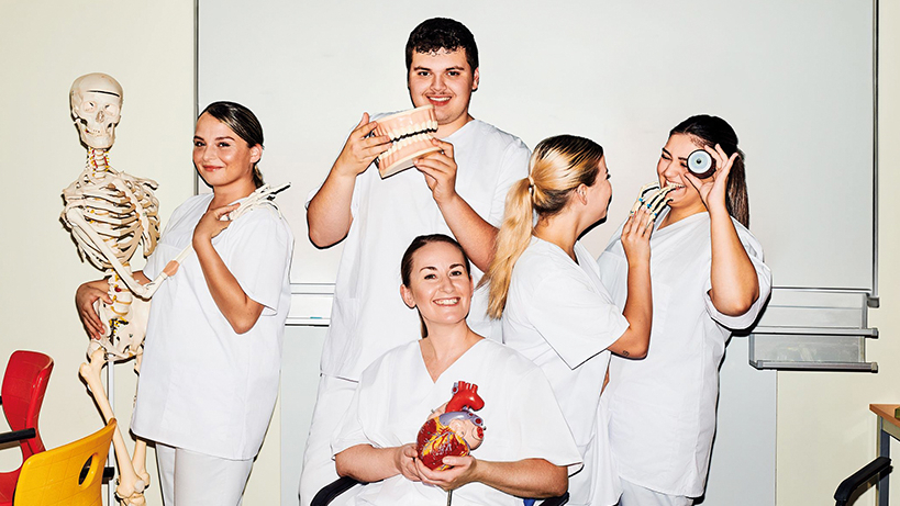 Fünf junge Menschen in weißer Pflegebekleidung mit Modellen von menschlichen Körperteilen und Organen