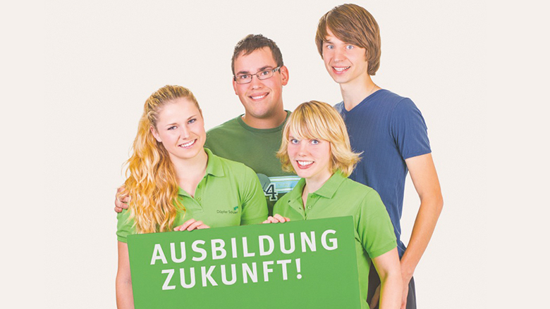 Vier Personen lächeln und halten ein grünes Schild mit der Aufschrift "AUSBILDUNG ZUKUNFT!" in die Kamera.