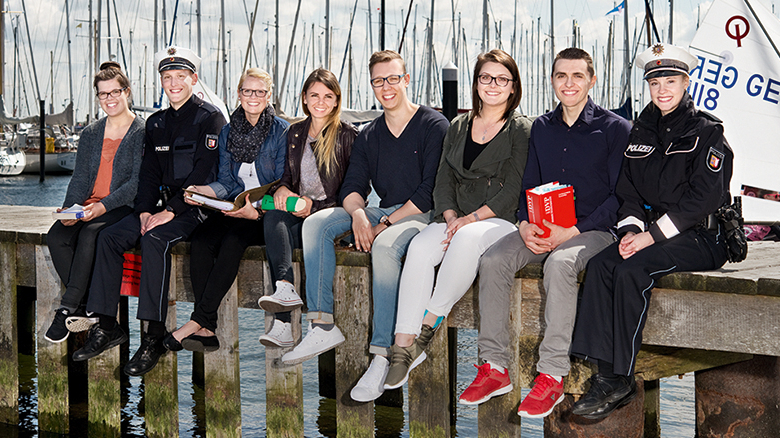 Acht Azubis aus verschiedenen Bereichen sitzen auf einem Steg, im Hintergrund Segelboote