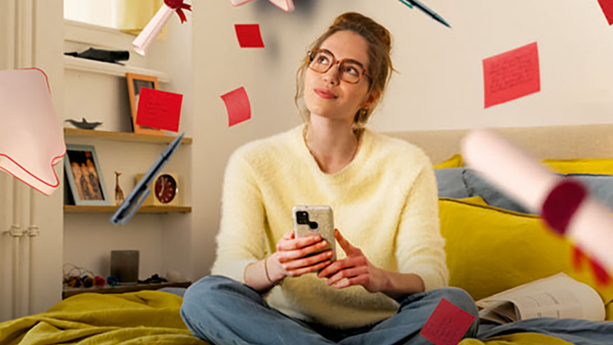 Eine junge Person sitzt mit einem Handy in der Hand in einem Zimmer auf einem Bett und blickt nachdenklich nach oben. Rote Zettel, Papierrollen und Stifte fliegen durch die Luft.