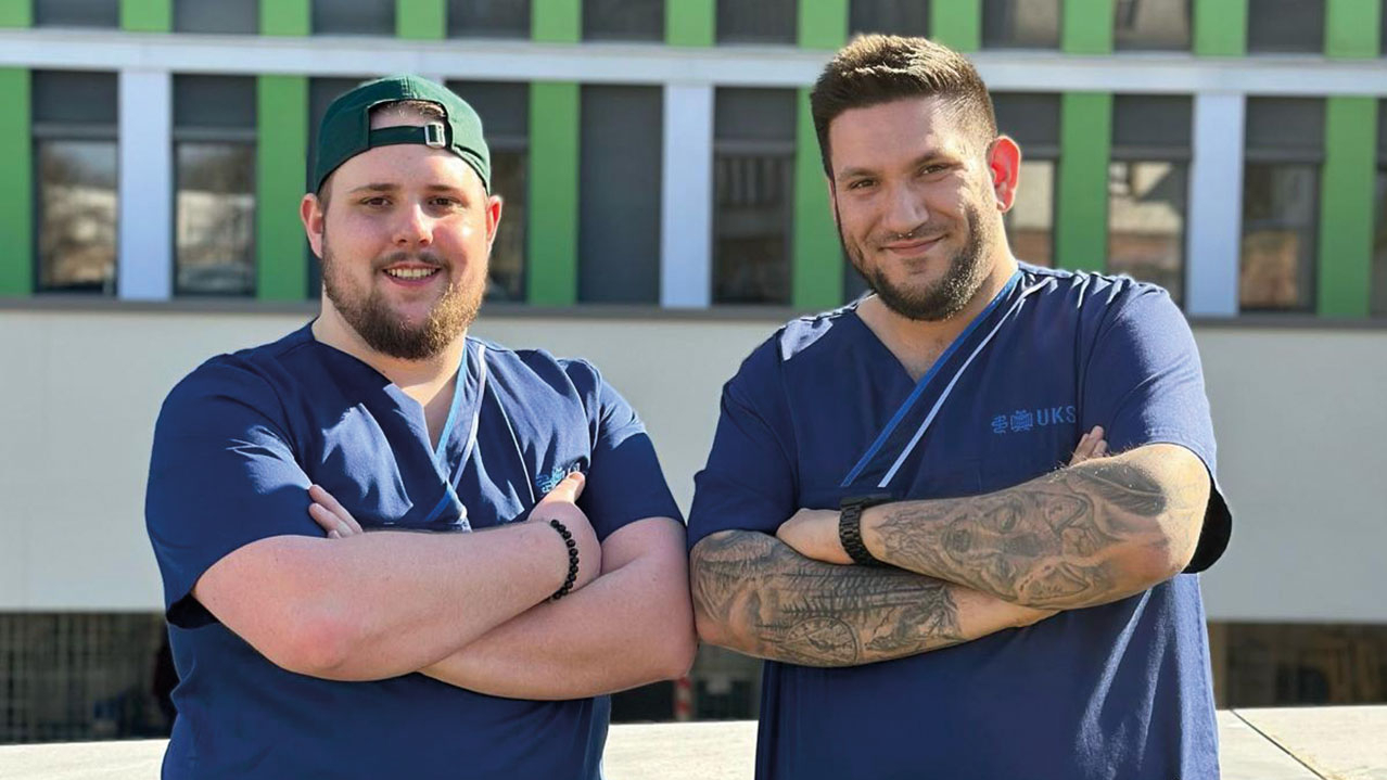 Zwei Pflegekräfte in blauen Kittel verschränken die Arme und lächeln