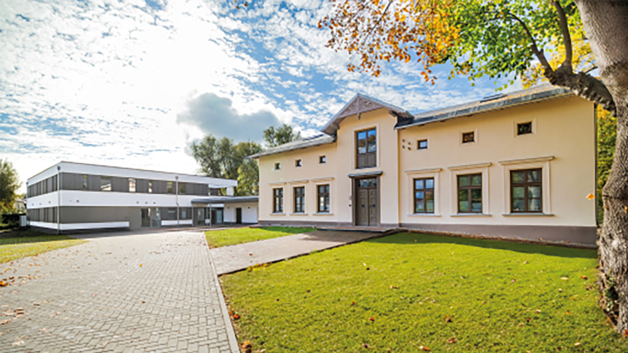 Gebäudeansicht mit Gehweg, Grünfläche und Laubbaum davor