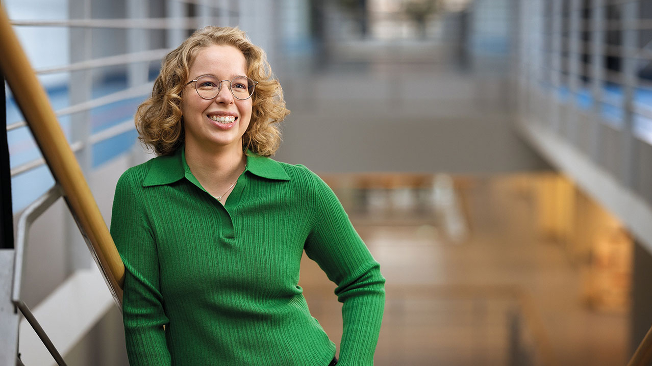 Junge Dame mit Brille und lockigem Haar in grünem Pullover steht auf einer Treppe und lehnt isch an das Geländer.