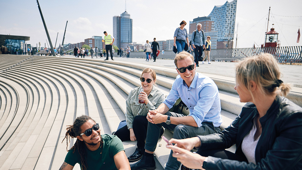 Vier junge Menschen sitzen auf einer Treppe an den Landungsbrücken, im Hintergrund die Elbphilharmonie