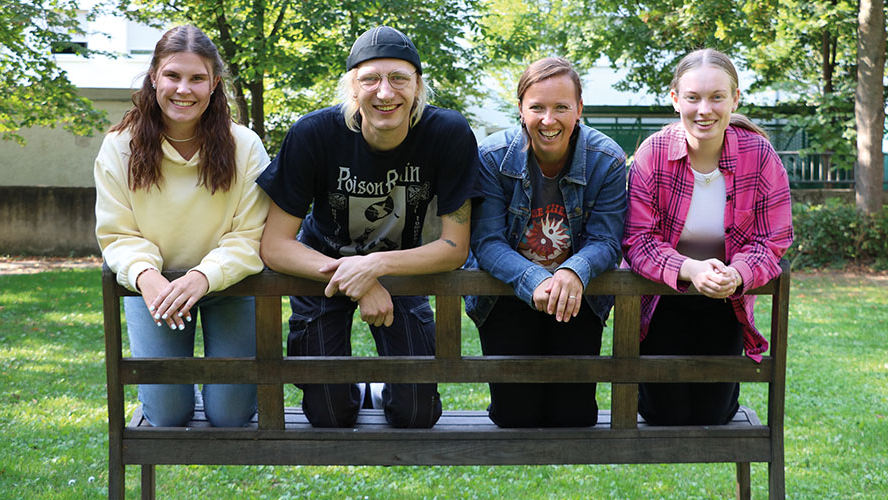 Vier Personen knien auf der Sitzfläche einer Bank und lächeln in die Kamera.