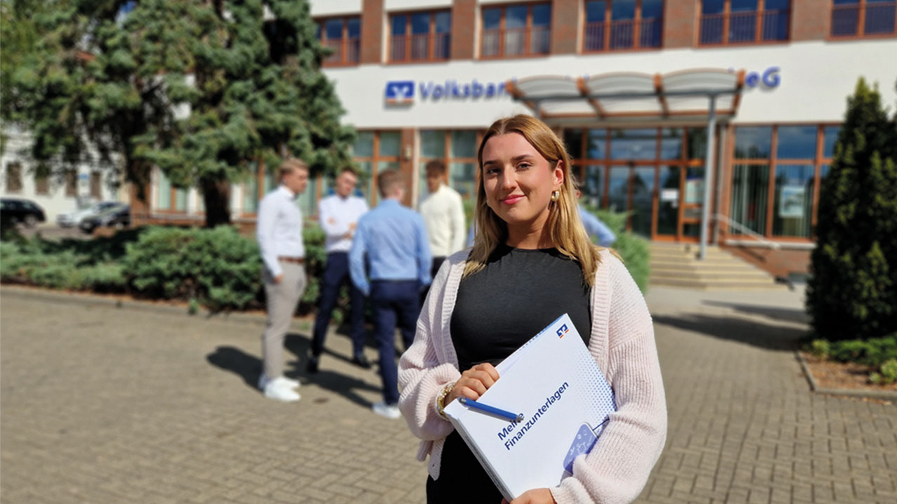 Junge weibliche Person mit Blonden Haaren hält eine Mappe in der Hand und steht vor einer Volksbank.