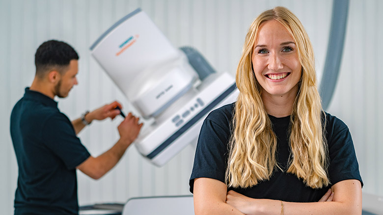 Zwei Personen, die Person im Vordergrund lächelt in die Kamera, die Person im Hintergrund arbeitet an einer Maschine.