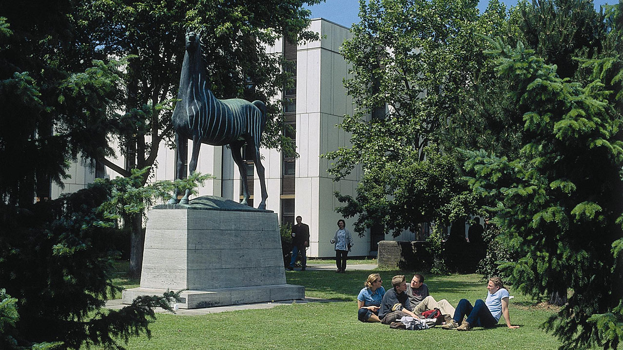 Vier Personen sitzen vor einer Statue im Park auf dem Boden. Im Hintergrund ist ein Gebäude sichtbar.
