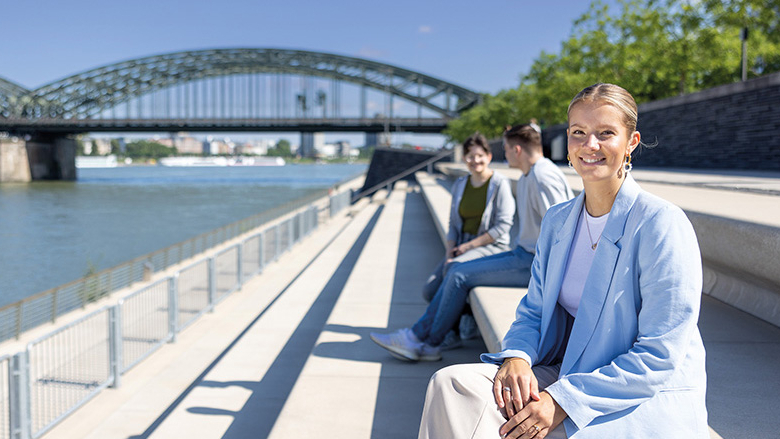 Junge Frau sitzt auf einer Steintreppe am Rhein und lächelt in die Kamera
