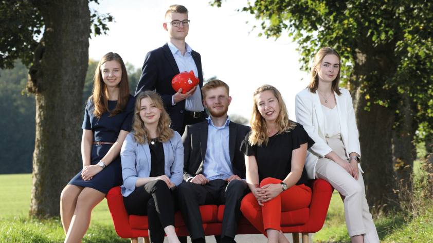 Fünf Personen sitzen auf einem roten Sofa im Grünen. Hinter dem Sofa steht eine Person mit einem roten Sparschwein in der Hand.