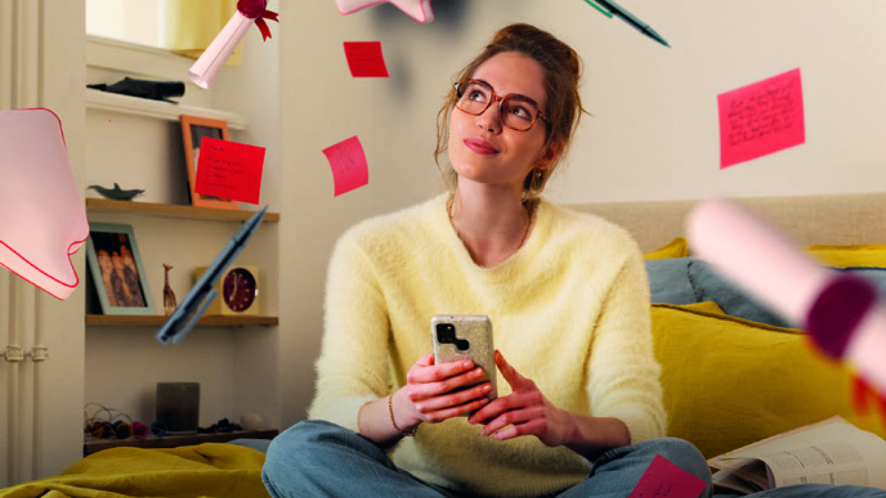 Werbekampagne: Junge Dame mit Brille sitzt auf einem Bett und viele Notizzettel fliegen durch das Zimmer.