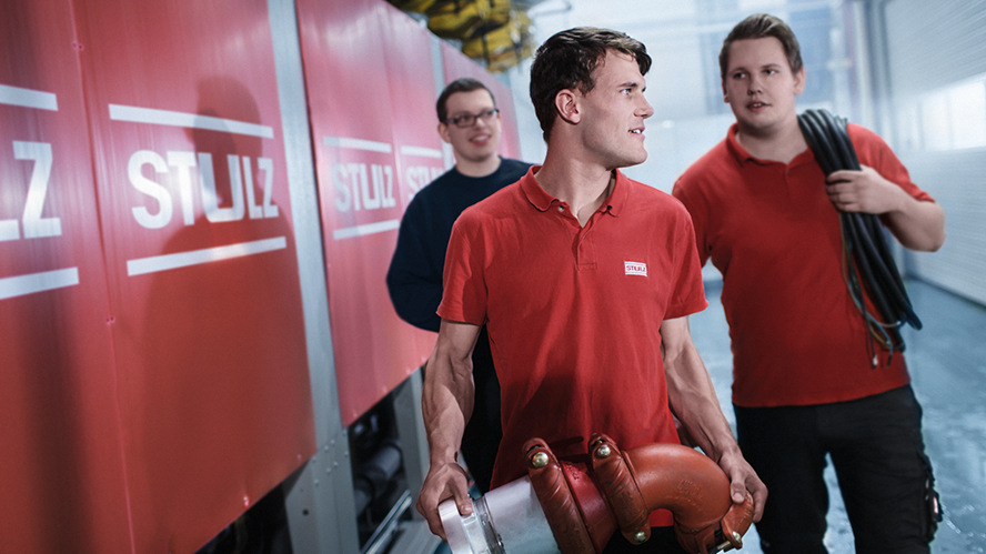 Drei junge Menschen gehen an einer Baustellenwand mit Firmenlogo entlang, einer davon trägt ein großes Rohrteil in der Hand