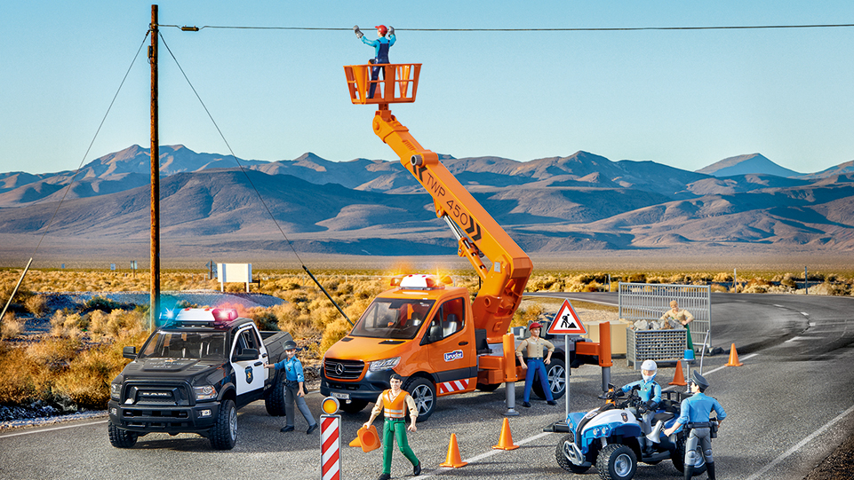 BRUDER Spielzeuge in einer Spielsituation: Auf einer Straße finden Arbeiten an der Oberleitung statt. Zwei Polizeifahrzeuge sperren die Straße ab, einige Bauarbeiter sind zu sehen.