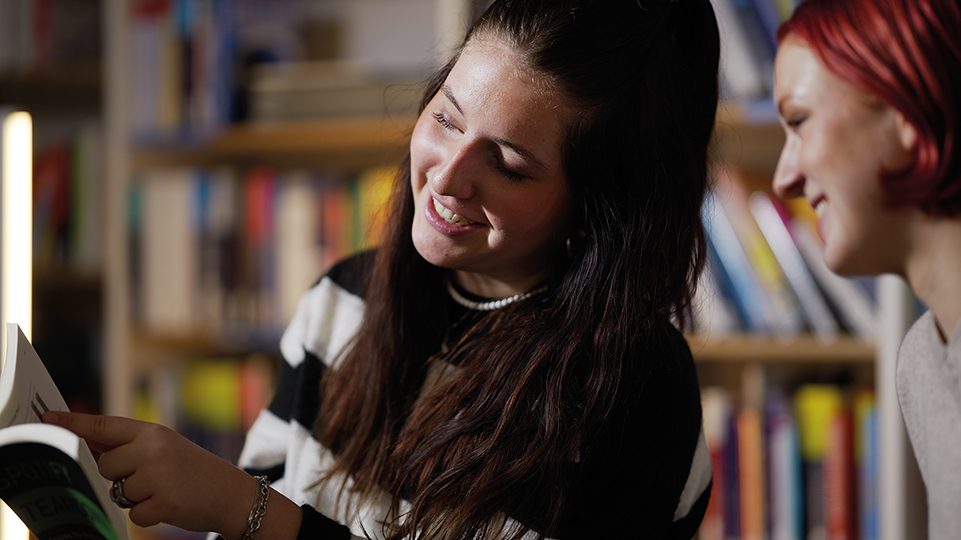 Zwei junge Frauen schauen vor Bücherwand gemeinsam in ein Buch