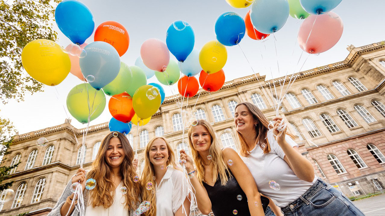 Vier lächelnde junge Damen stehen mit bunten Luftballons vor dem Universitätsgelände