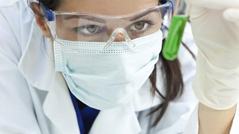 Großaufnahme einer jungen Frau mit Mundschutz und Schutzbrille, die sich ein Reagenzglas mit gründer Flüssigkeit ansieht