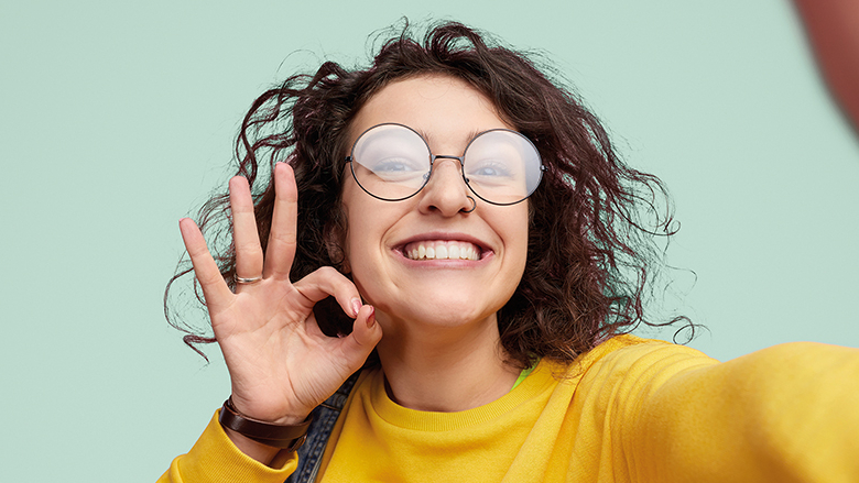 Junge Person mit Brille und lockigen Haaren formt mit Daumen und Zeigefinger das Zeichen "Exzellent" und lacht in die Kamera