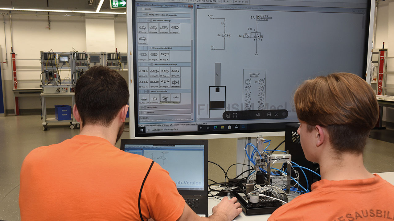 Zwei Personen in orangen Shirts sitzen an vor einem großen Bildschirm.