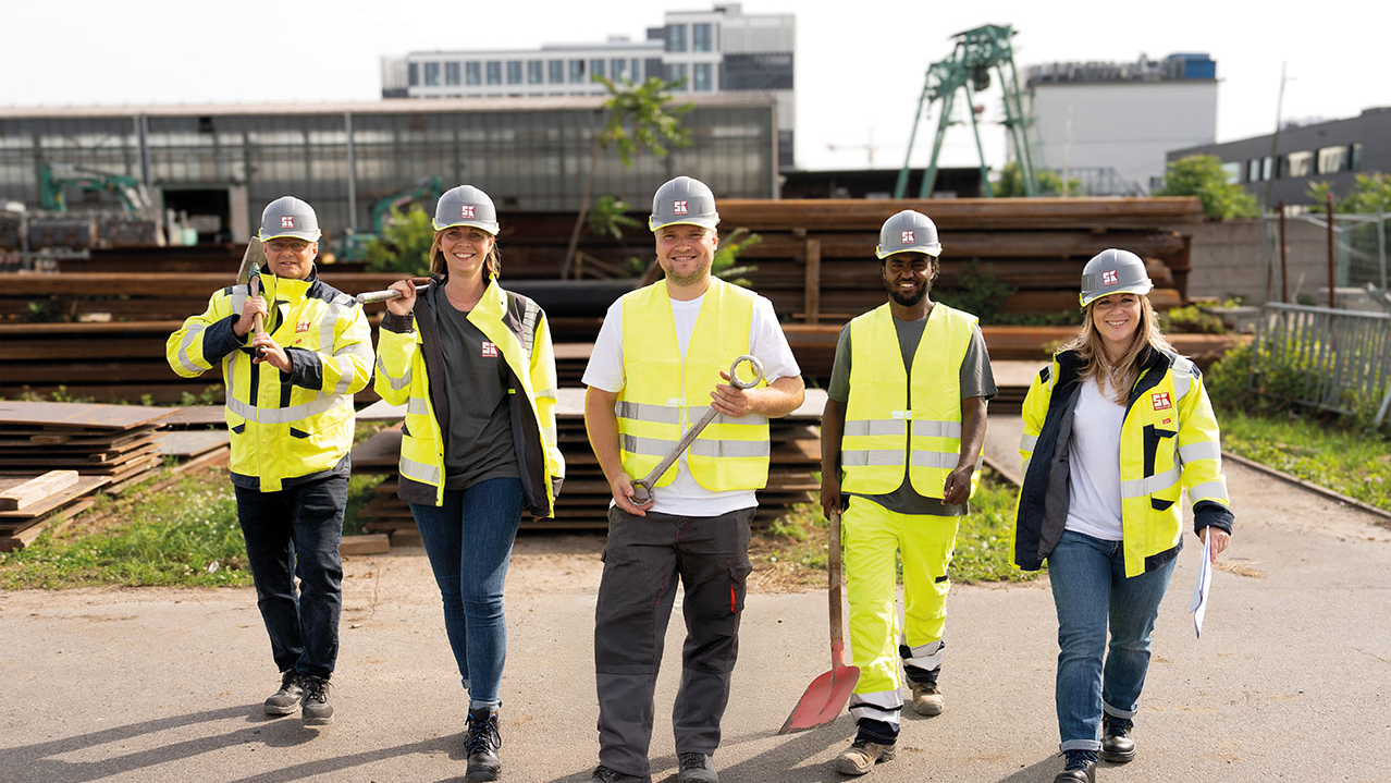 Personen in gelber Arbeitskleidung und Helm haben Schaufeln in der Hand. Sie stehen vor einer Industrieanlage mit Kran.