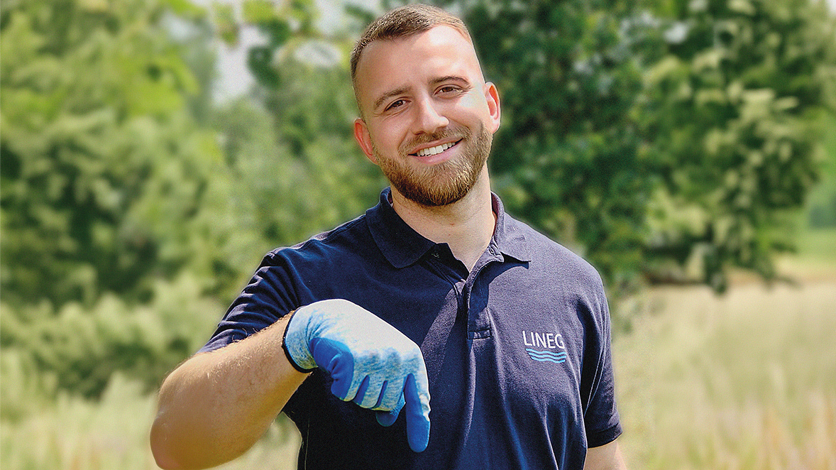 Junger Mann steht auf einem Feld und zeigt mit einer behandschuhter Hand nach unten