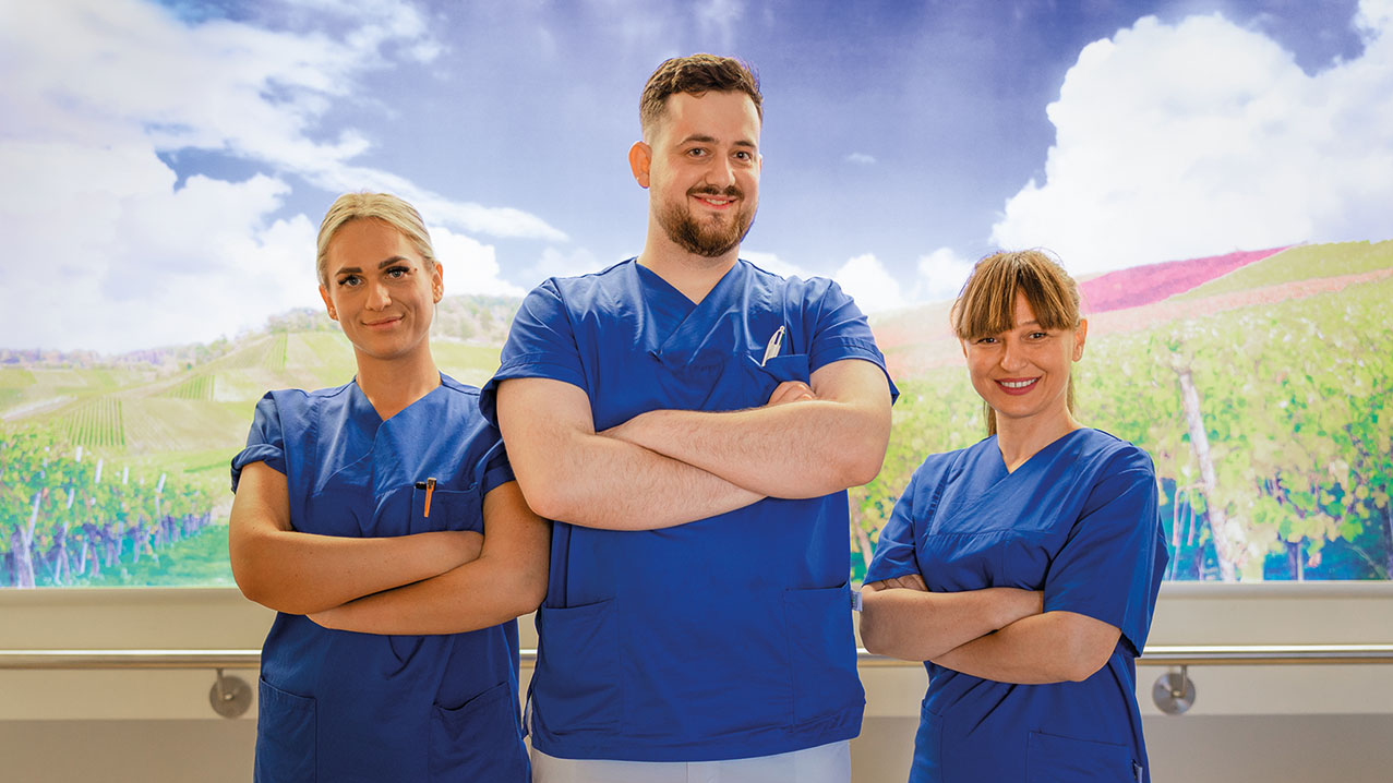 Drei medizinische Fachkräfte stehen nebeneinander, tragen blaue Kasacks und weiße Hosen, haben die Arme verschränkt und lächeln selbstbewusst. Im Hintergrund sind Weinberge zu sehen.