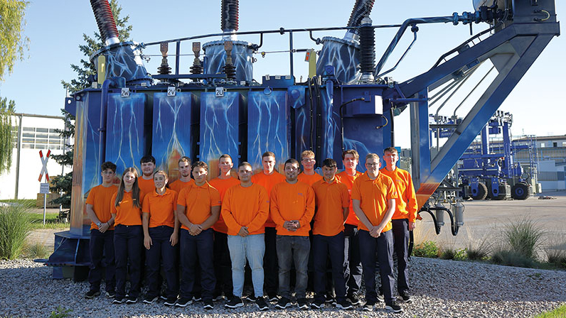 Fünfzehn Personen in oranger Kleidung stehen draußen vor einem großen, blau lackierten Industriegerät mit hohen Isolatoren.