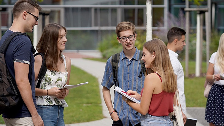 Zwei Gruppen Studierender vor einem Hochschulgebäude im lockeren Gespräch