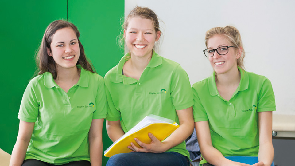 Drei junge Menschen in grünen Poloshirts mit Schullogo lächeln in die Kamera