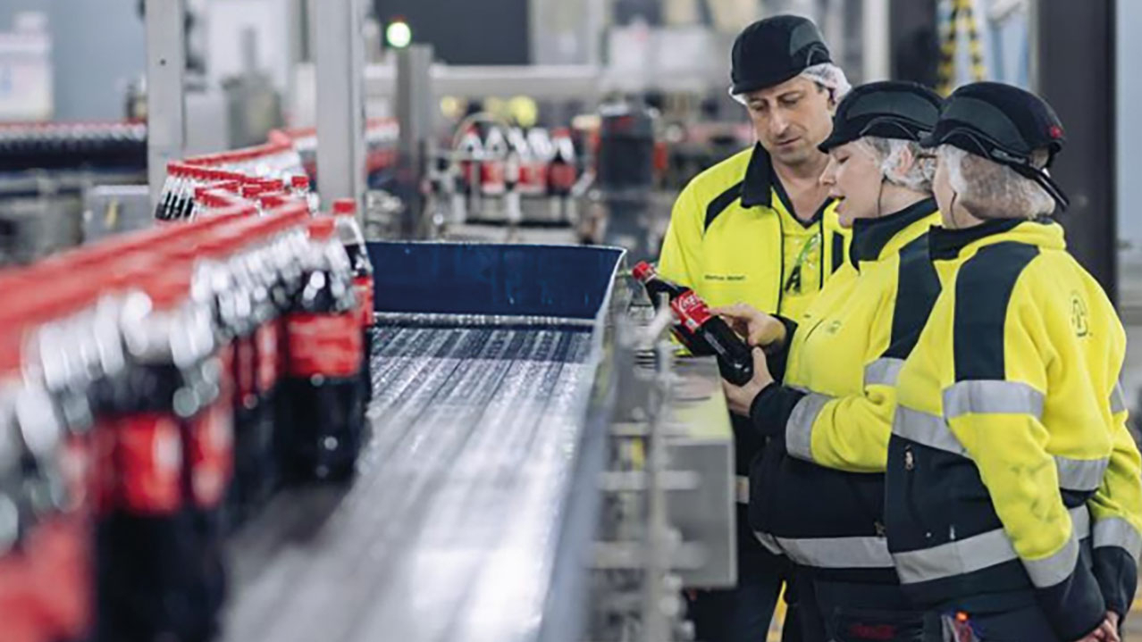 Mitarbeitende kontrollieren Coca Cola Flaschen am Fließband